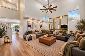 A spacious living room with a large sofa, a coffee table, and a ceiling fan. There is a staircase leading to an upper level, and a dining area is visible in the background.