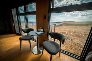 A room with a wooden floor, a round table with chairs, and a view of a beach and ocean through large windows.