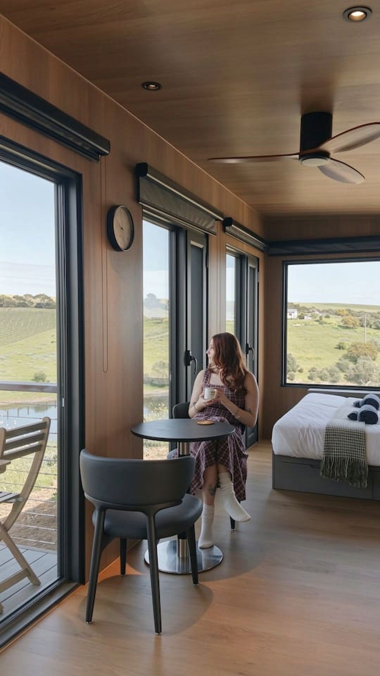 A room with a bed, a table, and a chair, large windows with a view of the countryside.