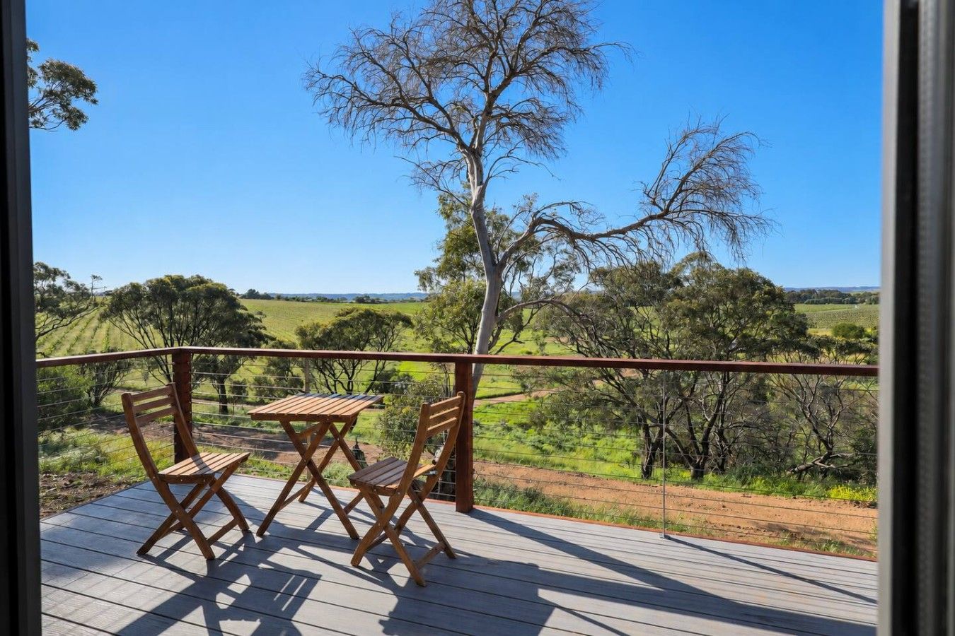 Een houten terras met een tafel en stoelen, met uitzicht op een landelijk landschap met bomen en velden.