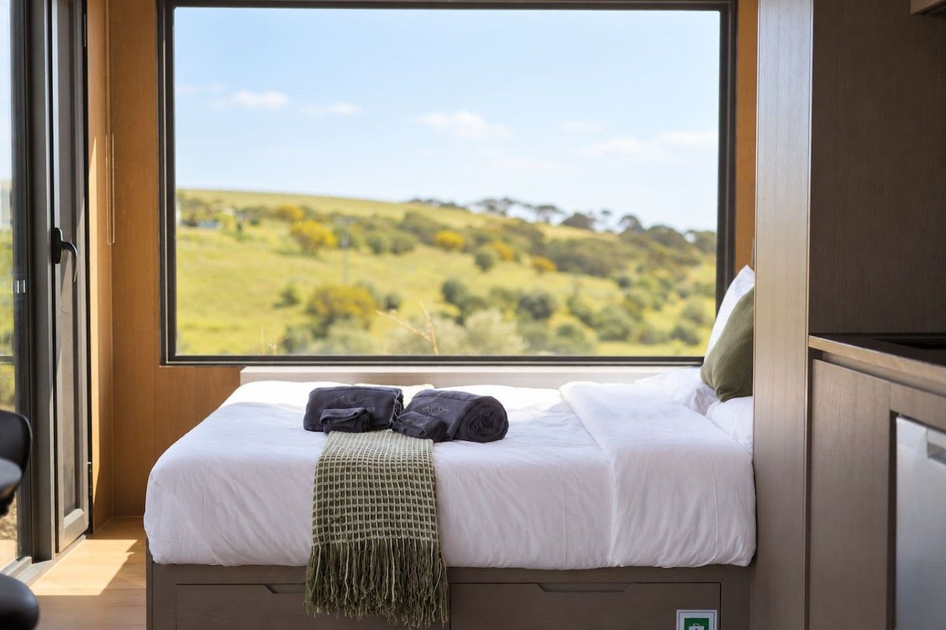 Een slaapkamer met een groot raam, een bed met wit beddengoed, twee donkere handdoeken en een groene plaid.