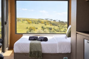 Een slaapkamer met een groot raam, een bed met wit beddengoed, twee donkere handdoeken en een groene plaid.
