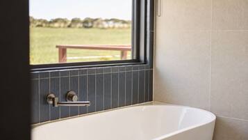 Een moderne badkamer met een vrijstaand bad, een raam met uitzicht op een grasveld en een betegelde muur.
