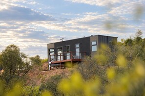 Een modern, twee verdiepingen hoog gebouw met een balkon, omgeven door natuurlijke begroeiing.