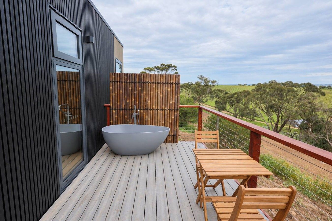 A modern outdoor deck with a large bathtub, wooden furniture, and a view of the countryside.