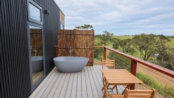 A modern outdoor deck with a large bathtub, wooden furniture, and a view of the countryside.