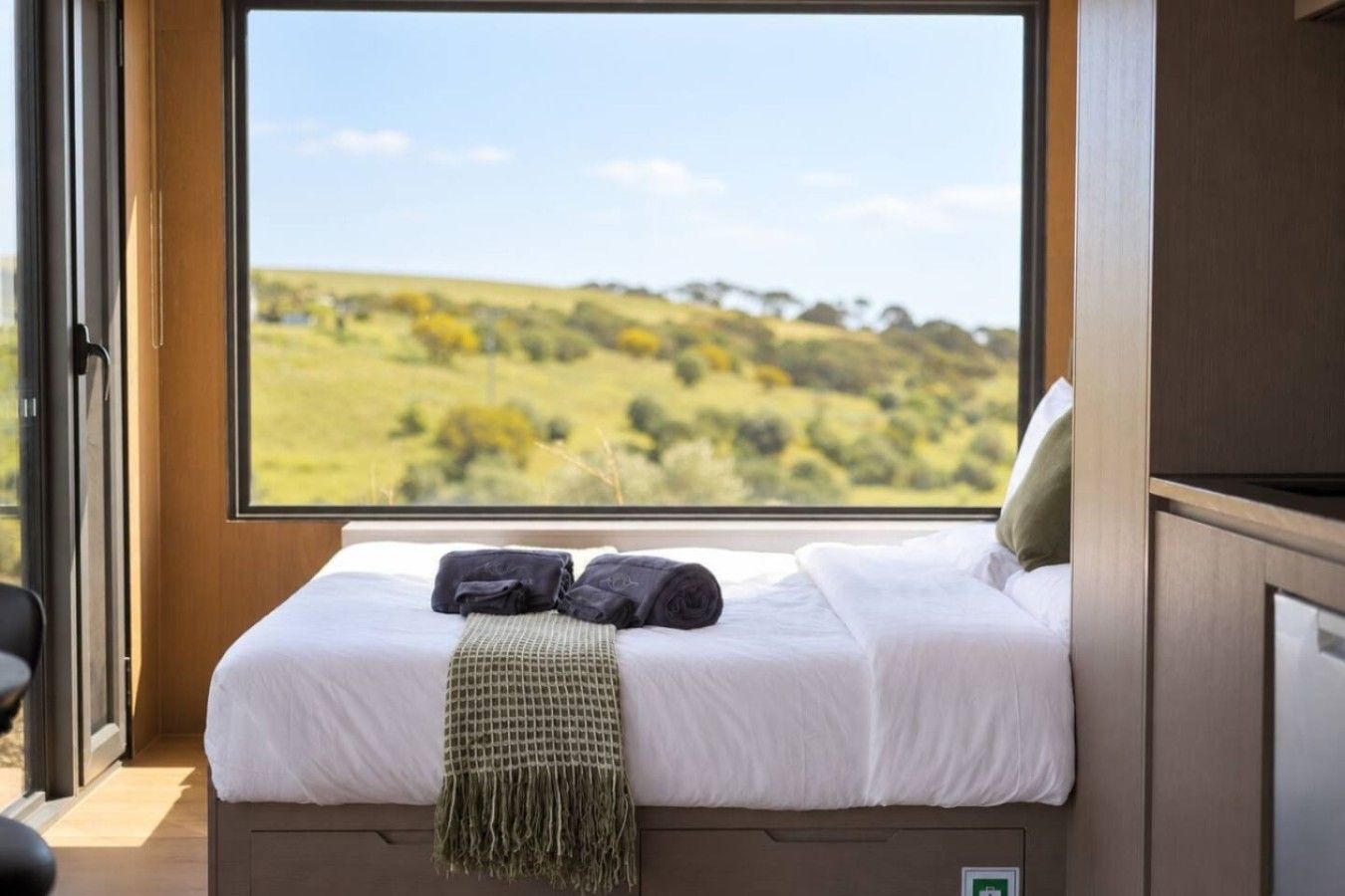 A bedroom with a large window, a bed with white bedding, two pillows, and a green throw blanket.