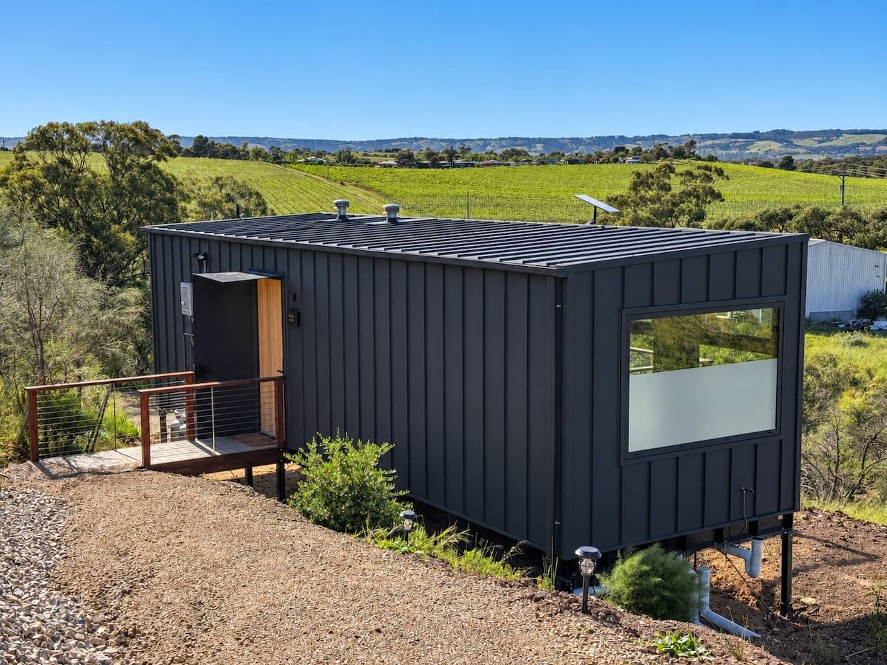 Une petite cabane aux couleurs sombres, dotée d’un petit porche et d’une grande fenêtre, située dans un paysage rural avec des collines ondulantes en arrière-plan.
