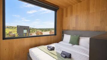 Une chambre moderne dotée d’une grande fenêtre, de murs en bois, d’un lit avec du linge de lit blanc et offrant une vue sur un paysage rural.