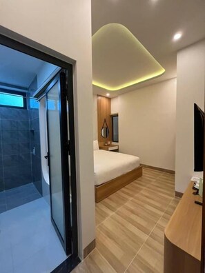 Ett modernt hotellrum med en glasdörr som leder till en duschhörna, en säng och ett skrivbord, alla tillverkade av trä.