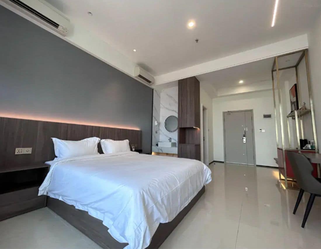 A modern hotel room with a large bed, a bedside table, a mirror, and a door leading to another room.