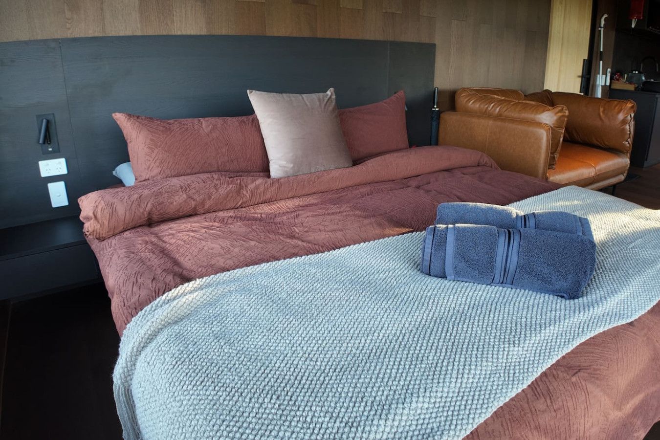 Un letto rifatto con una coperta e dei cuscini blu, un divano in pelle marrone e una testiera con un interruttore della luce.