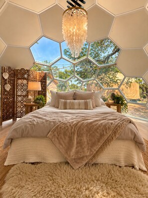 A bedroom with a hexagonal ceiling, a chandelier, a bed with multiple pillows, a wooden side table with a lamp, and a view of trees through the ceiling. - 6 bed resort Yurt at a luxury Austin Wellness  Ranch 10 minutes from downtown. (Austin)