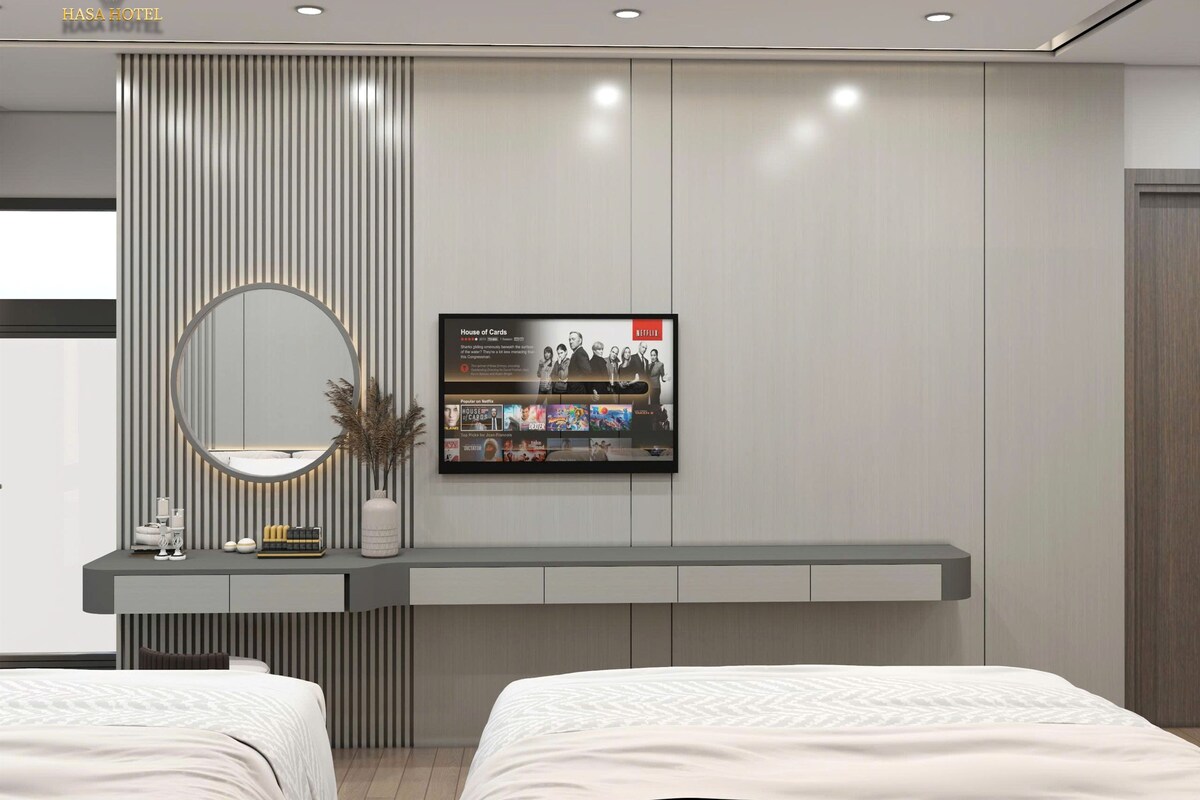 Quarto de hotel moderno com uma cama grande, mesa de cabeceira com espelho, TV fixada na parede exibindo uma série da Netflix e um vaso decorativo.