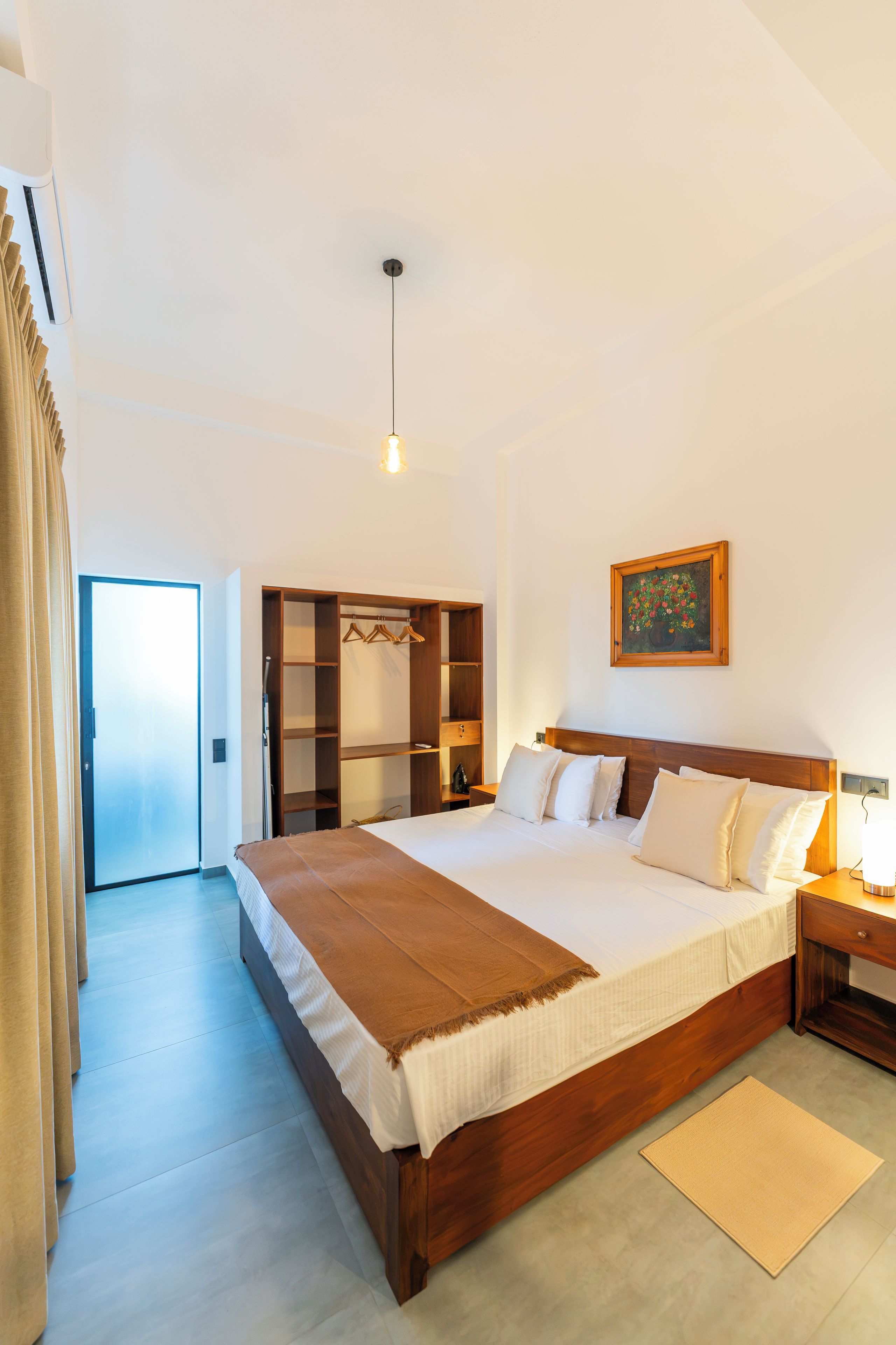 Habitación de hotel con una cama grande, cabecera de madera, mesita de noche con lámpara, armario con espacio para colgar y un cuadro en la pared.