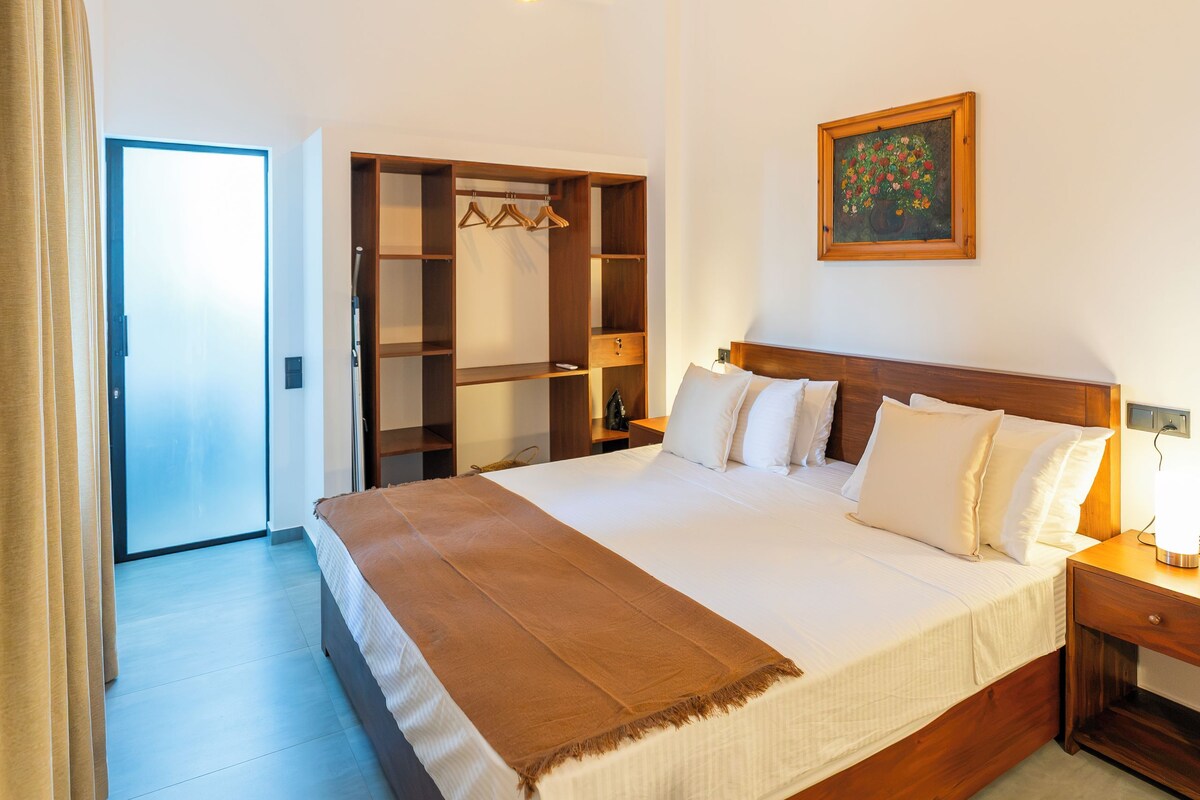 Una habitació d'hotel amb un llit gran, un capçal de fusta, una taula de nit amb una làmpada, un armari amb espai per penjar roba i un quadre a la paret.
