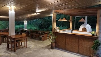 Zona d'aigües obertes amb taules i bancs de fusta, un taulell rústic de fusta amb un forn de pizza i llums penjants.