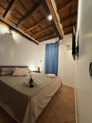 A bedroom with a wooden ceiling, a bed with a white bedspread, a bottle of champagne, and a blue curtain. - Bramante Collection – Fico Apartment By RHR (ROMA)
