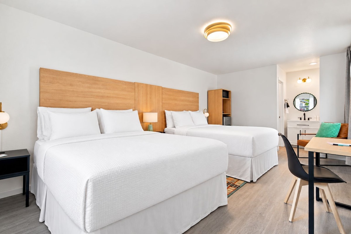 A hotel room with two beds, a desk, a chair, a nightstand, a lamp, a mirror, and a shelf.
