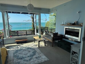 A living room with a large window overlooking the sea, a sofa, a coffee table, and a fireplace. - Shipwreck Bay beach house, surf and sunsets off the deck (Ahipara)