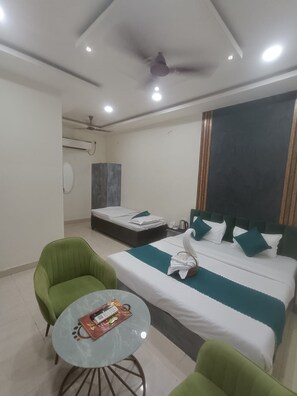 A hotel room with a bed, two green chairs, a small round table, a ceiling fan, and a wall-mounted air conditioning unit. - Radha Madhav Homestay (Varanasi)
