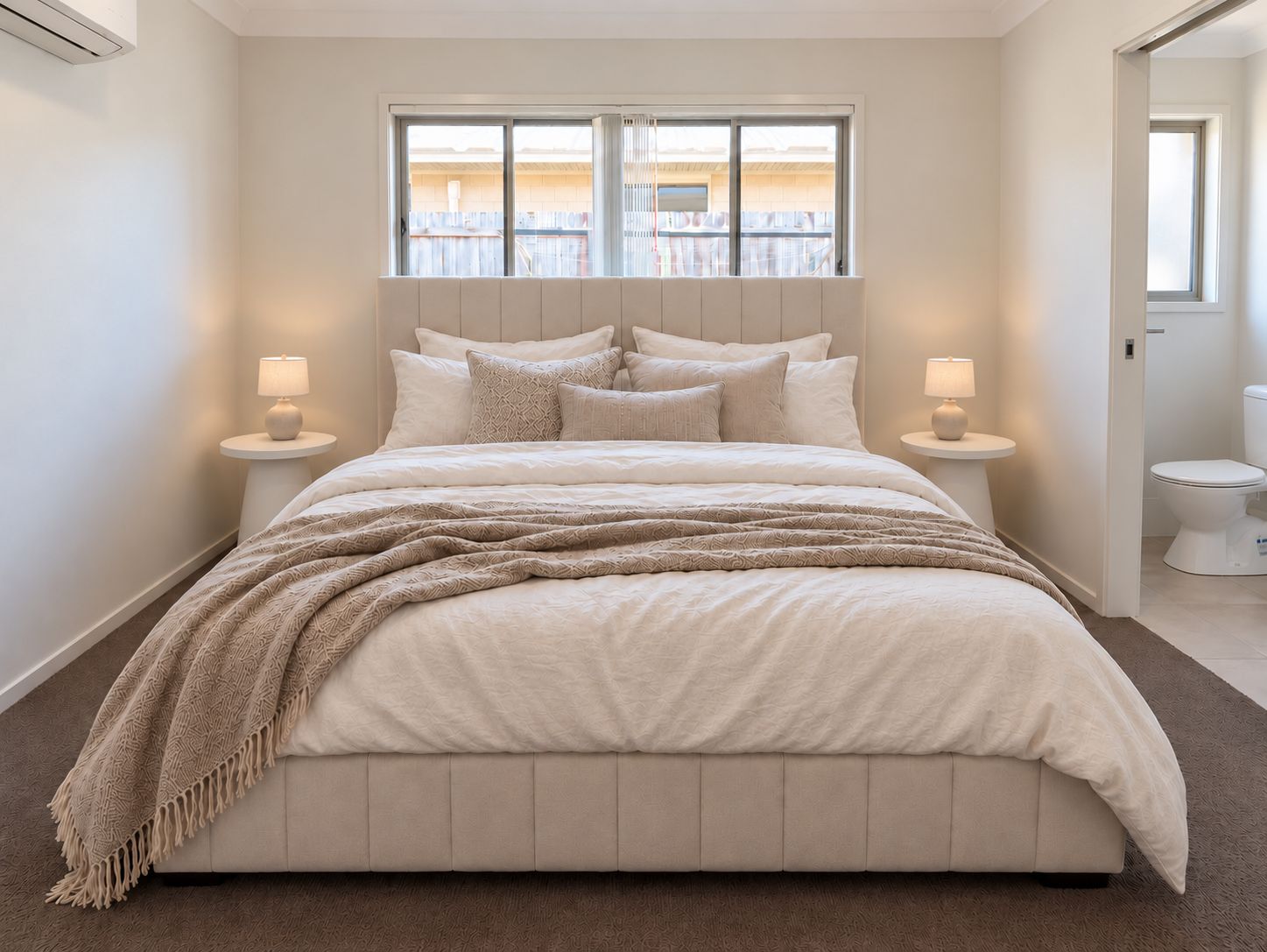 A bedroom with a large bed, two bedside tables with lamps, a window with blinds, and a view of the outside.