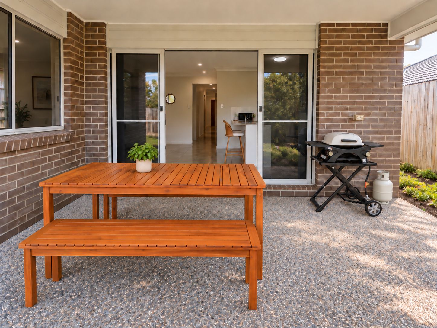 A patio with a wooden table and bench, a grill, and a potted plant.