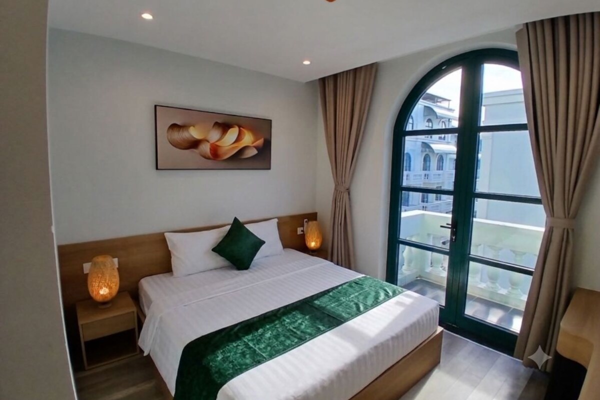 Een hotelkamer met een groot bed, een nachtkastje, een lamp, een schilderij aan de muur en een raam met gordijnen.
