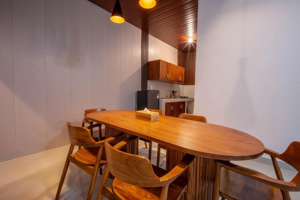 Un espace de salle à manger moderne avec une table et des chaises en bois, une cuisine en arrière-plan et des luminaires suspendus.