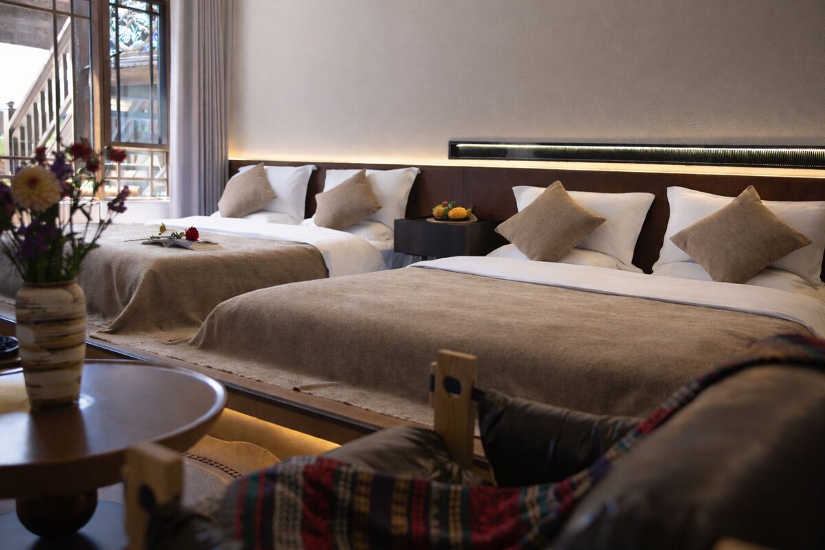 A hotel room with two beds, a nightstand, a vase with flowers, and a view of the street outside.