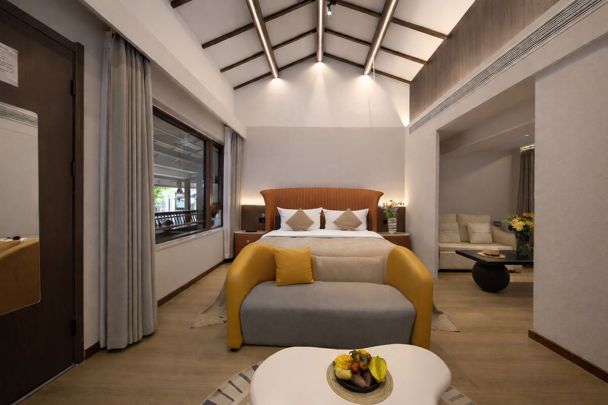 A modern hotel room with a large bed, a sofa, a dining table with a fruit bowl, and a view of the outdoors through a window.
