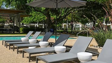 Area piscina con lettini e ombrelloni, circondata da vegetazione e un edificio sullo sfondo.
