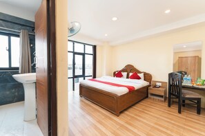 A hotel room with a wooden bed, a bedside table with a phone and water bottles, a desk with a chair, and a large window. - Pashupati Front Hotel (Kathmandu)