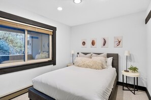 Ein modernes Schlafzimmer mit einem großen Bett, einem Fenster mit Jalousien, einem Nachttisch mit Lampe und Wandbildern.
