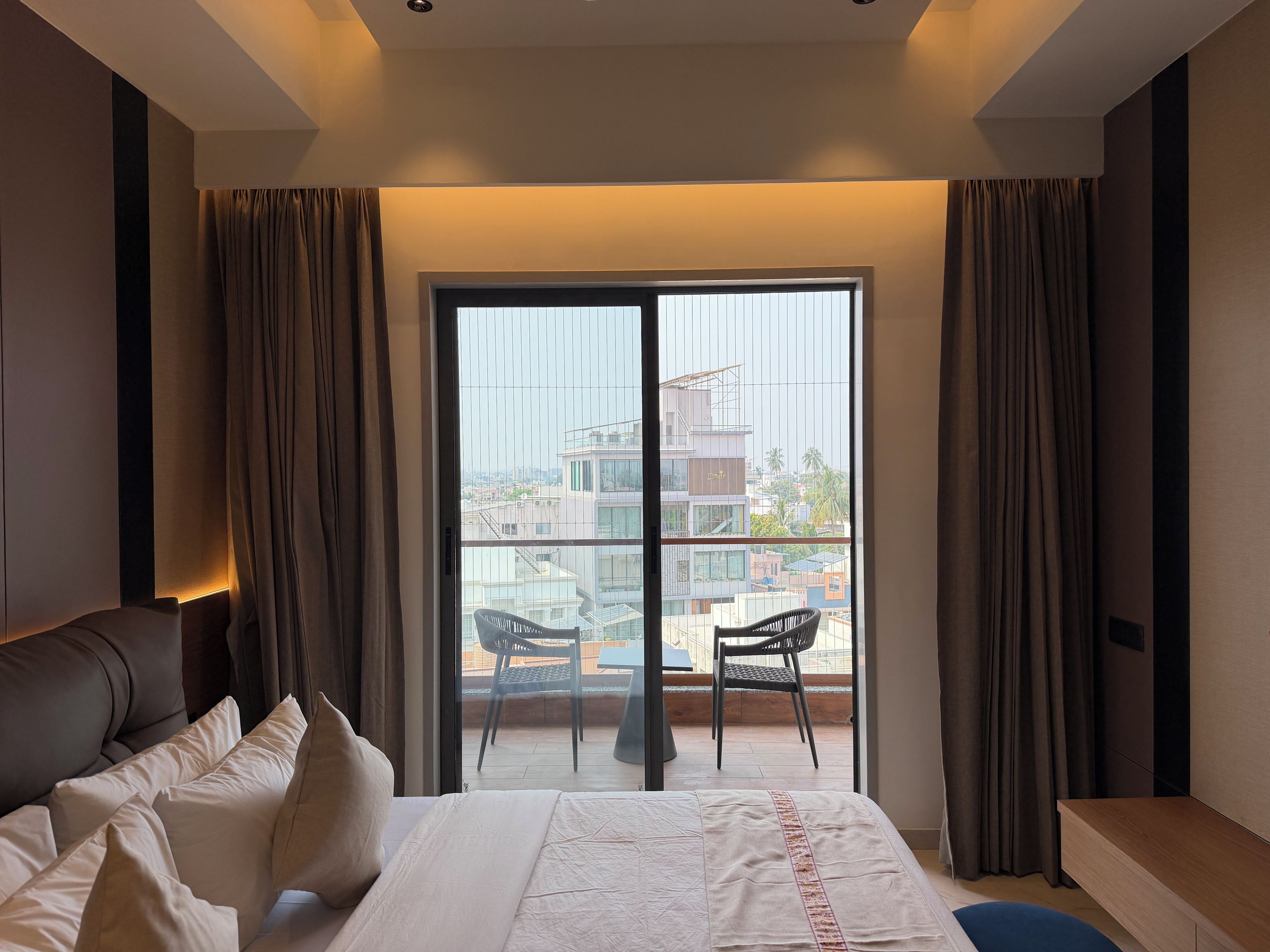 A hotel room with a bed, a desk, a chair, and a balcony with a view of buildings.