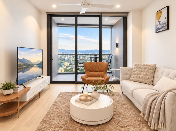Een moderne woonkamer met een grote televisie, een leren fauteuil, een witte bank en uitzicht op een stadsgezicht door grote ramen.