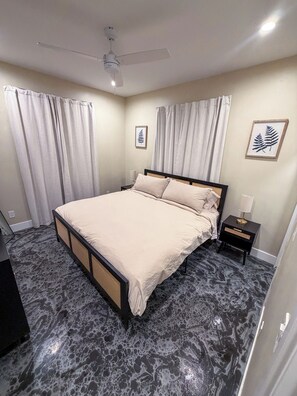 Une chambre à coucher comprenant un lit, une table de chevet, un ventilateur de plafond et deux décorations murales représentant des feuilles encadrées.
