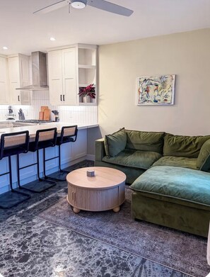 Un salon moderne avec un canapé vert, une table basse ronde et un coin cuisine doté de meubles blancs et d’un ventilateur de plafond.