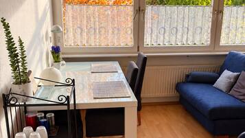Ein gemütliches Zimmer mit Esstisch, einer Couch, einem Bücherregal und Blick auf Herbstlaub durch die Fenster.