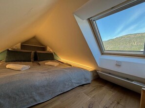 Een slaapkamer met een schuine kap, een bed met een grijze deken, een raam met uitzicht op een berg en een ingebouwde heater.