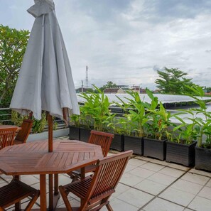 Una mesa de madera con sillas y un paraguas gris en un patio empedrado con plantas en macetas y vista a un cuerpo de agua.