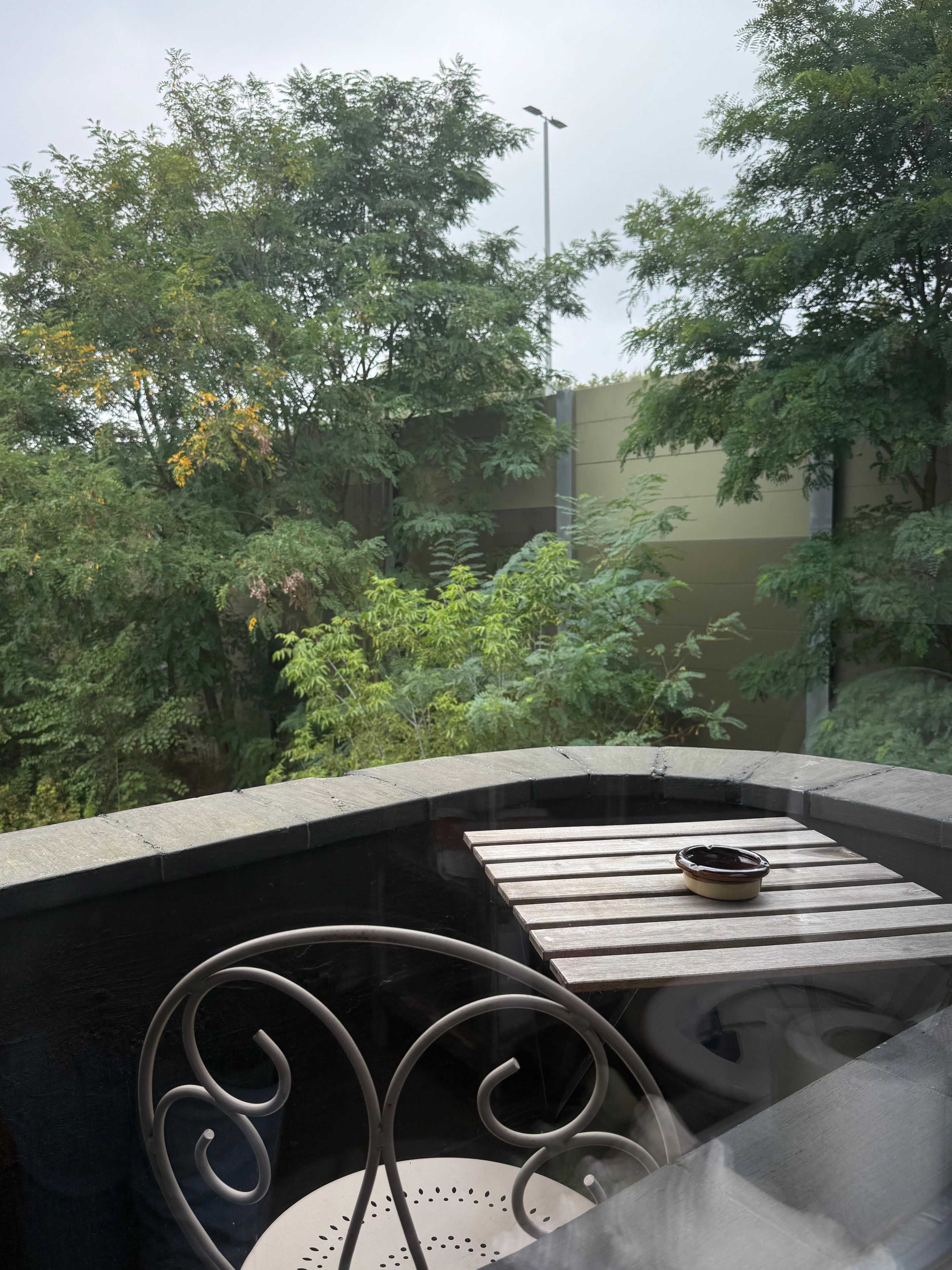 Un balcon avec une table et des chaises, donnant sur un jardin arboré et sur un bâtiment situé au loin.