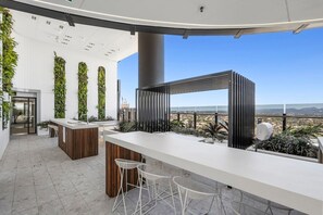 A modern bar area with a long counter, white bar stools, and a view of the cityscape. - Luxury 60th Floor | Panoramic Gold Coast Views | 2BR (Broadbeach)
