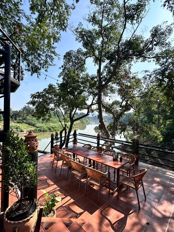 Un espacio de comedor al aire libre con una mesa y sillas, rodeado de plantas en macetas y vistas a un río.