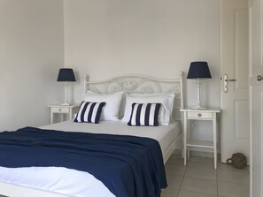 Una cama bien tendida con una colcha azul marino y almohadas blancas, flanqueada por mesitas de noche blancas con lámparas.