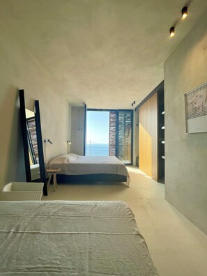 Une chambre à coucher moderne avec un grand lit, un petit tabouret et une vue sur la mer, visible à travers une porte coulissante.