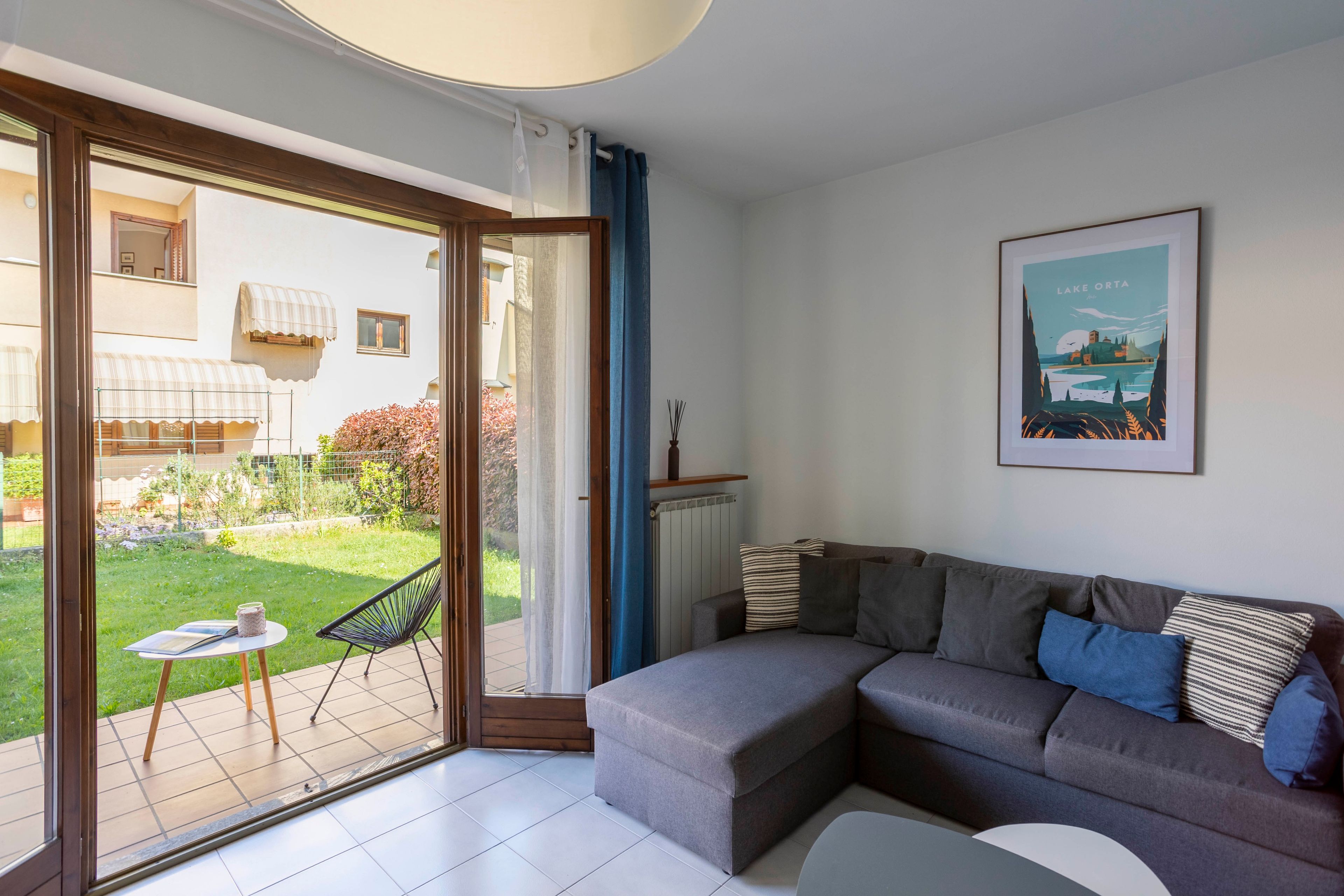 A modern living room with a grey sofa, a small round table, and a view of a garden through a sliding glass door.