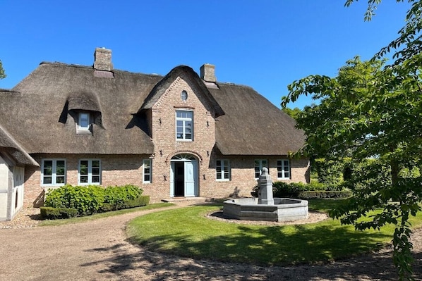 A traditional brick house with a thatched roof, a central entrance, and a small garden with a fountain.