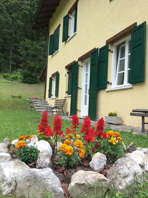 A yellow building with green shutters, a bench, and a flower bed with red and orange flowers. - Apartment '6 Posti' with Mountain View, Shared Garden and Wi-Fi (Borgo Valsugana)