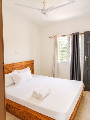 Una cama individual con cabecera de madera, ropa de cama blanca y dos toallas enrolladas. Se ve un ventilador de techo sobre la cama.